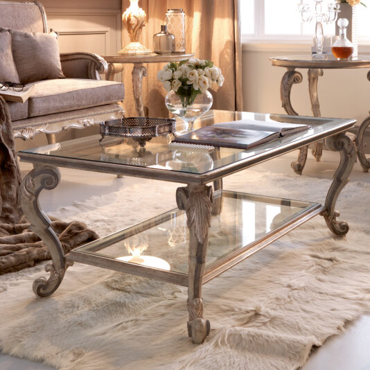 A luxurious living room features the Classic Italian Ornate Designer Glass Coffee Table, showcasing a glass top with intricately carved wooden legs and a lower glass shelf. The elegant table is adorned with a silver tray of decorative items and a vase of white flowers. Plush fur rugs and refined furniture enhance the sophisticated ambiance.