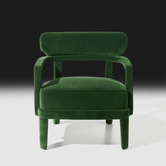 The Contemporary Small Velvet Armchair showcases deep green upholstery and a minimalist design. This modern chair is equipped with a cushioned seat and backrest, held by a robust rectangular frame with open sides and sleek four-legged support. The backdrop transitions from black at the top to white at the bottom, highlighting its stylish appeal.