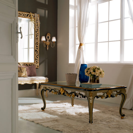 Ornate Carved Designer Italian Glass Coffee Table A luxurious room with an Ornate Carved Designer Italian Glass Coffee Table on a plush white rug, set near a large window with white drapes. In the background, a decorative mirror hangs above a cushioned bench, and a wall sconce with dual lamps adds subtle lighting.