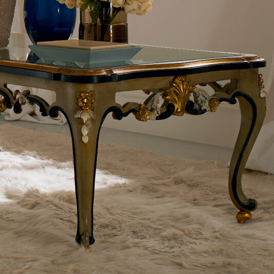 Ornate Carved Designer Italian Glass Coffee Table The Ornate Carved Designer Italian Glass Coffee Table, adorned with intricate carvings and gold accents, stands on a plush white rug. The table showcases decorative motifs, with a vase of flowers and several other items placed on top. The setting exudes a luxurious ambiance.