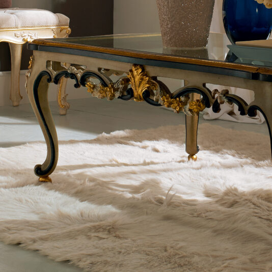 Ornate Carved Designer Italian Glass Coffee Table This image showcases the Ornate Carved Designer Italian Glass Coffee Table with gold accents. It stands on a plush, white fur rug, next to beige furniture pieces and a large decorative vase in the background. The room exudes a luxurious and elegant decor style.