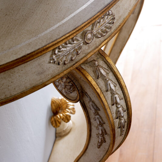Close-up of an ornate leg from the Designer Italian Demi Lune Console Table, showcasing intricate carvings and gold accents. The curved design with detailed leaf patterns is painted in a distressed white finish with gold highlights, and it is elegantly set on a wooden floor.