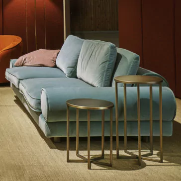 A cozy living room featuring a plush, light blue Contemporary Retro Inspired Designer Velvet Sofa with a pink pillow. In front of the sofa are two modern round nesting tables with thin metal legs. The room has muted lighting, beige carpeting, and a warm, dark red wall in the background, creating a contemporary retro-inspired ambiance.