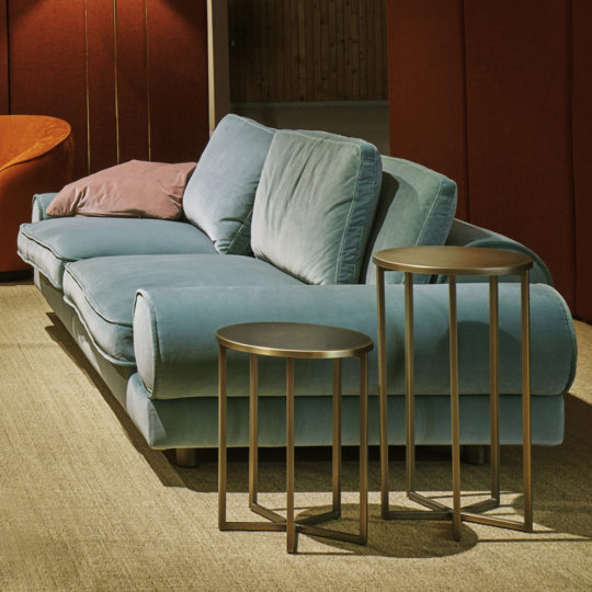 A cozy living room featuring a plush, light blue Contemporary Retro Inspired Designer Velvet Sofa with a pink pillow. In front of the sofa are two modern round nesting tables with thin metal legs. The room has muted lighting, beige carpeting, and a warm, dark red wall in the background, creating a contemporary retro-inspired ambiance.