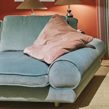 A Contemporary Retro Inspired Designer Velvet Sofa in teal is placed against a red wall. It features comfortable, rounded armrests with a soft, beige pillow partially leaning on one of them. Behind the sofa are two matching designer table lamps, and to the right hang striped curtains. The floor is carpeted in beige.