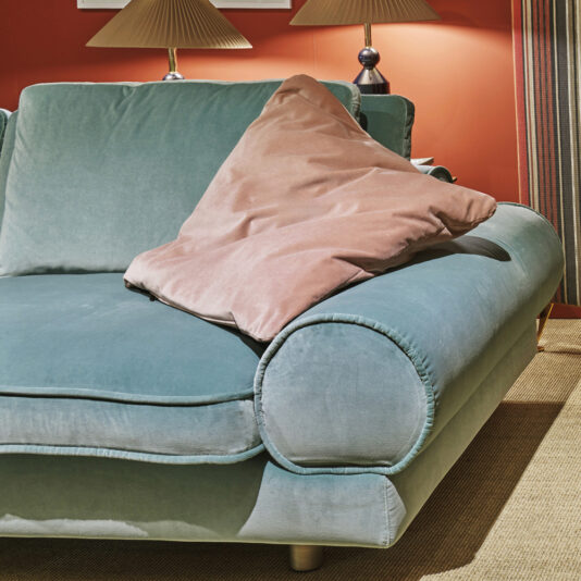 Contemporary Retro Inspired Designer Velvet Sofa A Contemporary Retro Inspired Designer Velvet Sofa in teal is placed against a red wall. It features comfortable, rounded armrests with a soft, beige pillow partially leaning on one of them. Behind the sofa are two matching designer table lamps, and to the right hang striped curtains. The floor is carpeted in beige.
