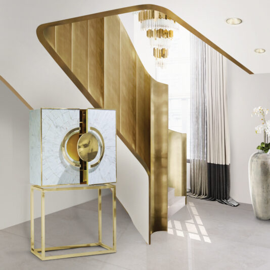 Exclusive Marble Art Deco Inspired Contemporary Cabinet A modern interior features a geometric gold staircase with a suspended chandelier. To the left of the staircase stands the Exclusive Marble Art Deco Inspired Contemporary Cabinet, characterized by its shattered-glass effect and gold accents, adding a touch of elegance. Natural light streams in through a large window adorned with white and gray curtains.
