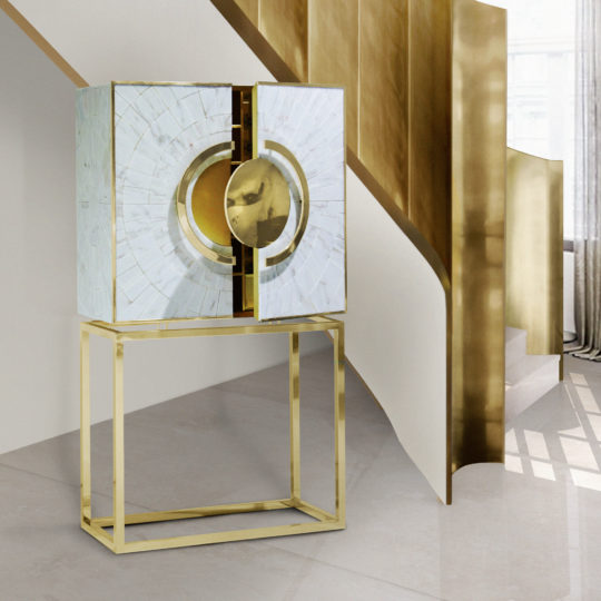The Exclusive Marble Art Deco Inspired Contemporary Cabinet, with its geometric design, stands open against a large staircase. This modern piece features a circular, gold center and a mosaic of pale, reflective panels. Supported by a sleek golden frame, it complements the shadowed wall and stairs in the background.