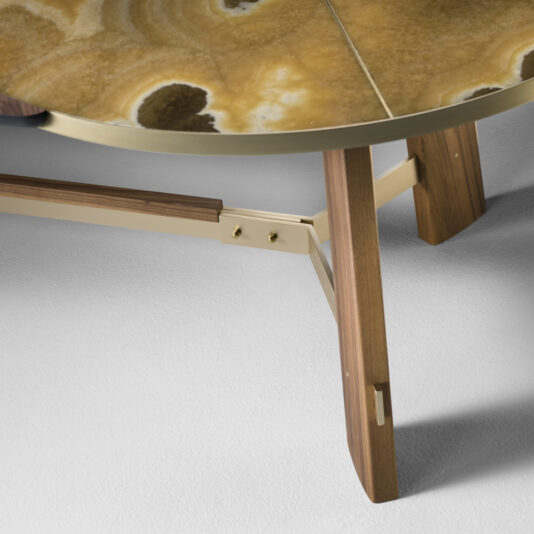 Close-up of the Oval Walnut And Marble Contemporary Designer Dining Table, featuring a distinctive brown and yellow marbled top with sleek, gold-colored metal bracing and wooden legs with a natural finish. The table's sophisticated design is showcased on a light, textured surface.