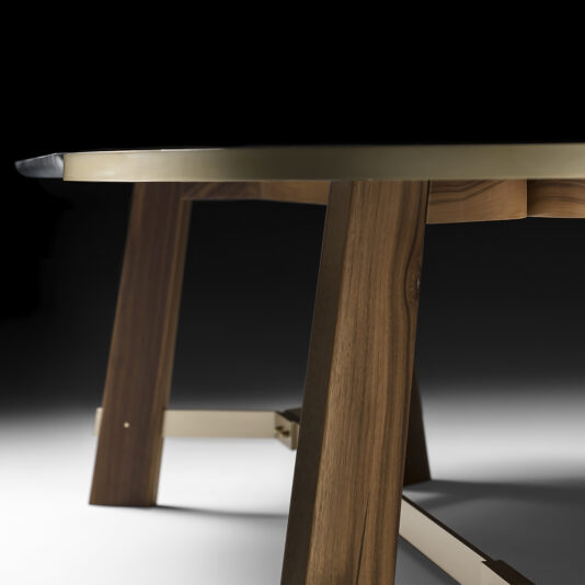 A close-up angled shot of the Oval Walnut And Marble Contemporary Designer Dining Table with a sleek design. The table features a smooth, rounded edge and sturdy legs reinforced by metal accents. The dark background emphasizes the table's elegant structure and finish.