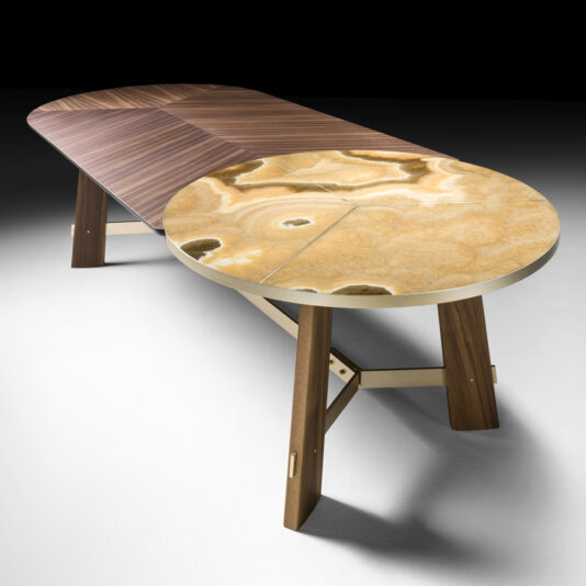 The Oval Walnut And Marble Contemporary Designer Dining Table is a sleek, modern piece that features a unique fusion of materials: one half with a smooth, striped wooden surface and the other half covered in polished, abstract-patterned stone. It stands on wooden legs complemented by metal supports.