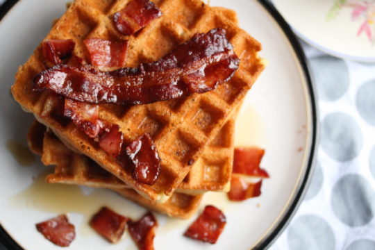 A plate of golden-brown waffles topped with crispy bacon slices and drizzled with syrup.