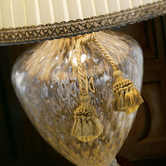 A close-up of the Luxury Engraved Hand Blown Murano Glass Table Lamp showcases its intricately patterned base, adorned with gold tassels. The beige lampshade, trimmed with dark lace, creates a vintage yet elegant look. The softly illuminated background hints at wood furniture.
