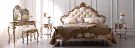 An elegant bedroom featuring a luxurious bed with an ornate, tufted headboard, flanked by matching nightstands adorned with floral arrangements. A vanity set with a large mirror and cushioned stool stands to the left, all showcasing sophisticated, classic styling reminiscent of luxury Italian furniture.