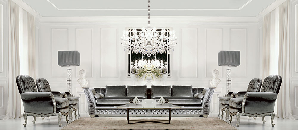 A luxurious, modern living room features a gray velvet sofa, matching armchairs, and a glass coffee table. Adorned with luxury Italian furniture, the room boasts a large crystal chandelier, two gray floor lamps, and elegant white paneling on the walls, complemented by a centerpiece of white flowers.