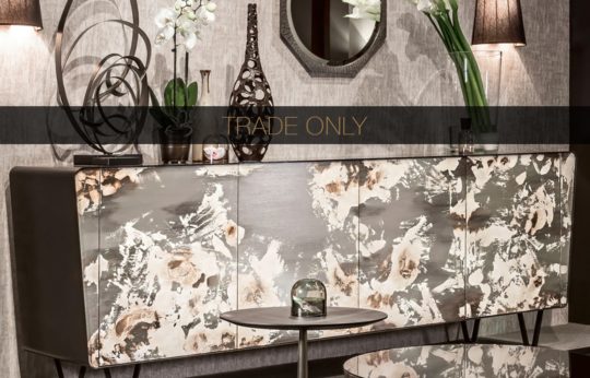 A modern living room setting featuring a stylish sideboard with abstract-patterned doors. Decorative items, including a vase, sculpture, and orchid plant, adorn the top of the sideboard. A round mirror and two tall lamps are mounted on the wall above. Text overlay reads "TRADE ONLY.