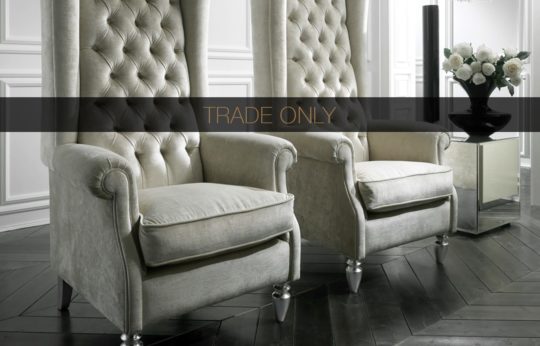 Two elegant, light grey, tufted high-back armchairs are positioned side-by-side on a dark, herringbone-patterned wooden floor. A black vase with white flowers sits on a mirrored side table next to the chairs. The overlay text reads "TRADE ONLY.