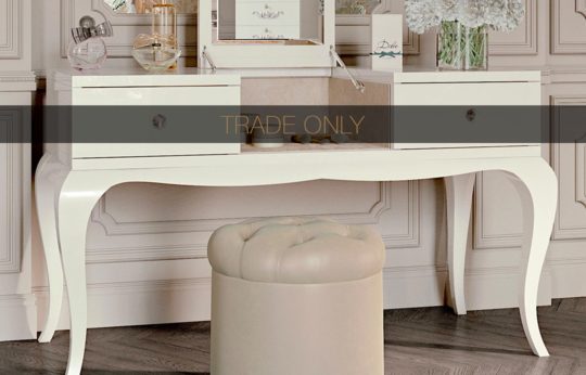A white vanity table with an open central compartment, flanked by two closed drawers, stands against a wall with intricate paneling. A cushioned beige stool sits in front, and a decorative vase with flowers is on top. The words "TRADE ONLY" overlay the image.