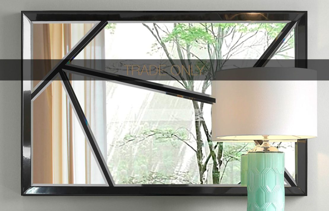 A modern mirror with an abstract geometric black frame is mounted on a wall. A light-colored curtain and tree are reflected in the mirror. A blue ceramic lamp with a white shade sits on a table in front of it. The words "TRADE ONLY" are overlaid on the mirror.