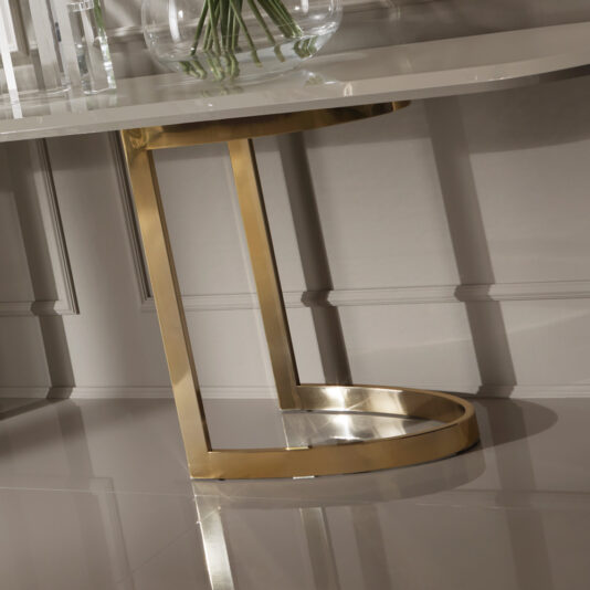24 Carat Gold Plated Oval Designer Console Table Close-up of the 24 Carat Gold Plated Oval Designer Console Table showcasing its beige top and striking gold-colored metal base. The geometric base features a circular bottom and vertical supports, set against a decorative paneled wall.