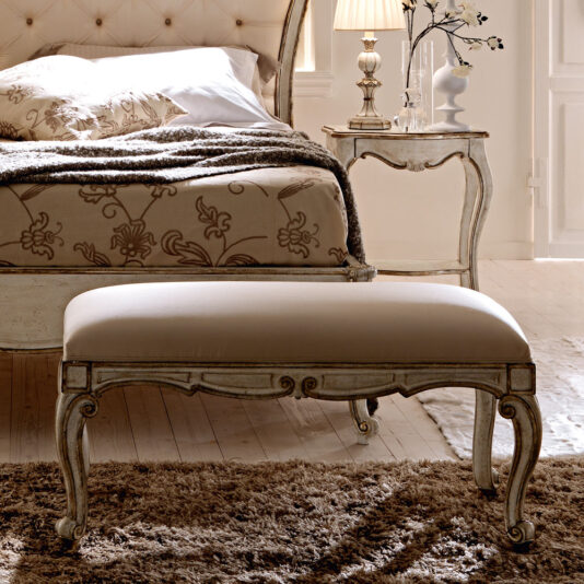 An exquisitely plush, beige upholstered small bench from the Antiqued White Louis Designer collection, featuring ornate, carved wooden legs, rests on a fluffy brown rug in a luxurious vintage-style bedroom. The room is complemented by an antiqued white Louis designer bed frame adorned with patterned bedding and an elegant nightstand decorated with a traditional lamp and various decorative items.