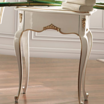 A White Louis Reproduction Bedside Table stands on a polished wooden floor. The table's design features elegant, curved legs and decorative carvings with gold accents. Stacks of books rest on top, with part of a lamp visible in the background.