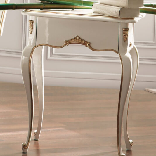 White Louis Reproduction Bedside Table A White Louis Reproduction Bedside Table stands on a polished wooden floor. The table's design features elegant, curved legs and decorative carvings with gold accents. Stacks of books rest on top, with part of a lamp visible in the background.