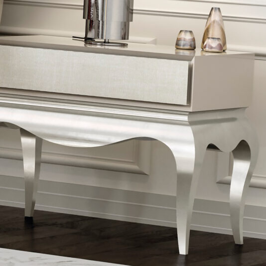 Luxury Silver Leaf Bedside Table A sophisticated metallic table with curved legs stands against a wall with wainscoting. The tabletop features a reflective decorative piece and two small candle holders, evoking the elegance of the Luxury Silver Leaf Bedside Table and enhancing the room's refined atmosphere.