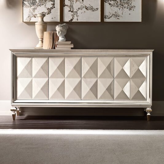 The Italian Diamond Design Mother of Pearl Sideboard features a stylish Italian diamond pattern on its doors, complemented by light wood finishes and decorative details. The top is adorned with books, a classical-style bust, and vases, while abstract framed artworks provide a captivating backdrop on the wall behind it.