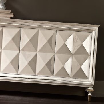 Italian Diamond Design Mother of Pearl Sideboard A close-up of the Italian Diamond Design Mother of Pearl Sideboard showcases its elegant front panels adorned with a geometric, pyramid-patterned design. The piece features a glossy finish and delicate, angled legs, standing against a dark, solid-colored wall. Its mother of pearl accents add a touch of opulence to the overall aesthetic.