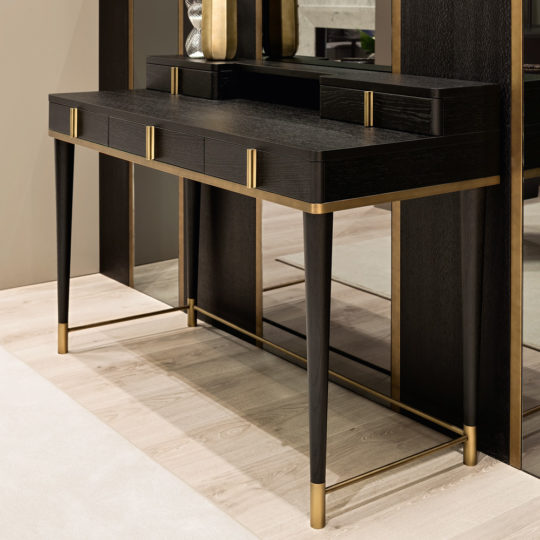 The Black Oak Italian Designer Writing Desk, featuring gold accents and tapered legs, stands on a light wood floor against a mirrored wall with decorative vases placed on top.