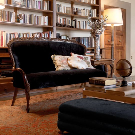 A cozy library with tall bookshelves, a Classic Italian Designer Walnut Day Sofa adorned with decorative pillows, a wooden coffee table with books and a globe, a patterned rug, and warm floor lamp lighting.