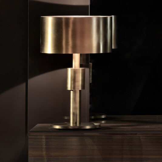 The Contemporary Burnished Brass Luxury Table Lamp, featuring a cylindrical shade and base, stands on a dark wooden surface against a dark background, casting soft light and creating a subtle reflection.