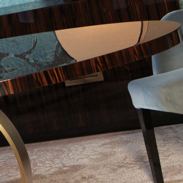 Contemporary Italian Bronzed Ring Oval Dining Table A partial view of a modern room featuring a Contemporary Italian Bronzed Ring Oval Dining Table with a reflective surface, accompanied by a cushioned chair upholstered in light blue. The background showcases a dark, patterned wall, while the floor is covered by a soft, textured carpet.