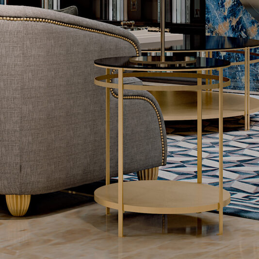 A modern living room corner features a grey upholstered sofa with gold nailhead trim, a Contemporary Italian Gold Round Lamp Table with a sleek glass surface, and a geometric patterned rug on a polished floor. A blue marble accent adds an elegant touch in the background.