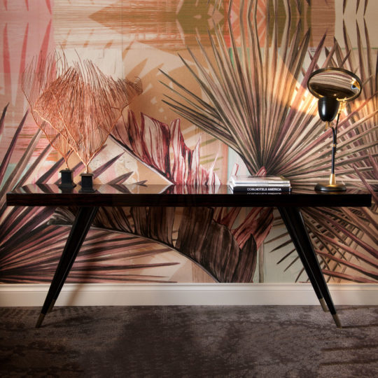 A Contemporary Italian Rectangular Console Table displays a gold lamp, two dried palm leaves in black vases, and stacked books, set before wallpaper featuring large tropical leaf patterns in earthy tones.