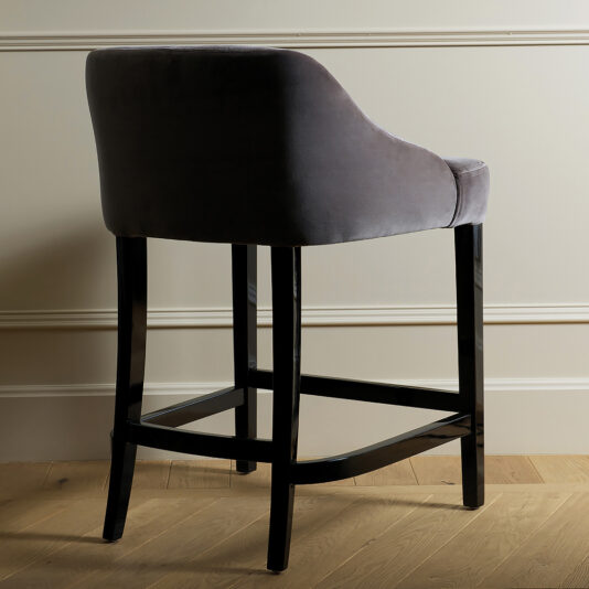 The Contemporary Italian Upholstered Velvet Bar Chair showcases a sleek and modern design with a dark grey velvet seat and backrest. Supported by four black wooden legs, this bar chair features a curved back and footrest, standing elegantly on a wooden floor against a light-colored wall with decorative molding.
