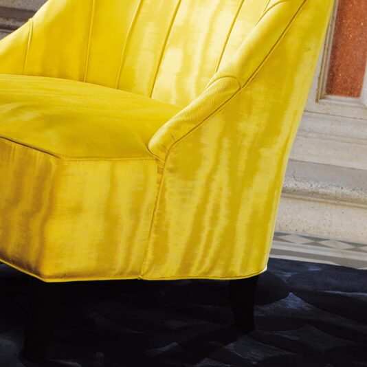 A close-up of the Contemporary Luxury Italian Armchair, upholstered in luxurious bright yellow velvet. The chair showcases a ribbed backrest and stands on dark wooden legs, positioned on a black patterned rug against the backdrop of stone steps and a partial view of a reddish-brown wall.