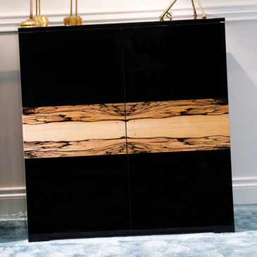 Contemporary Luxury Italian Cabinet The Contemporary Luxury Italian Cabinet, characterized by its sophisticated black finish and a unique horizontal wooden stripe with a natural, textured pattern across the front, stands elegantly against a light grey wall. The lower portion is settled on a blue carpeted floor, with decorative items thoughtfully placed on top.