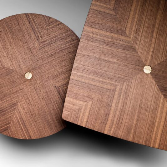 Contemporary Walnut Designer Side Table Close-up of two Contemporary Walnut Designer Side Tables showcasing geometric designs on their wooden tabletops. The circular tabletop exhibits a radiating pattern, and the square tabletop mirrors this design. Both tables feature a small, round metal accent at the center and are crafted from rich walnut wood. They are positioned at an angle against a neutral backdrop.