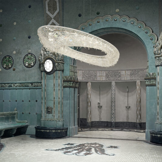 A luxurious, ornate room with teal and green accents features a Large Modern Cut Crystal Chandelier. The walls and columns are adorned with intricate patterns and mosaic tiles. In the background, two shower niches are set into an arched alcove with intricate wall designs.