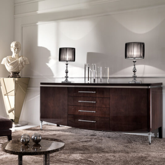 A modern living room showcases a Designer High Gloss Walnut Veneer Buffet, adorned with two elegant black lamps featuring pleated shades and a collection of clear glass vases. Next to the sideboard stands a statue on a pedestal, accompanied by metallic spherical decor on an adjacent small table.