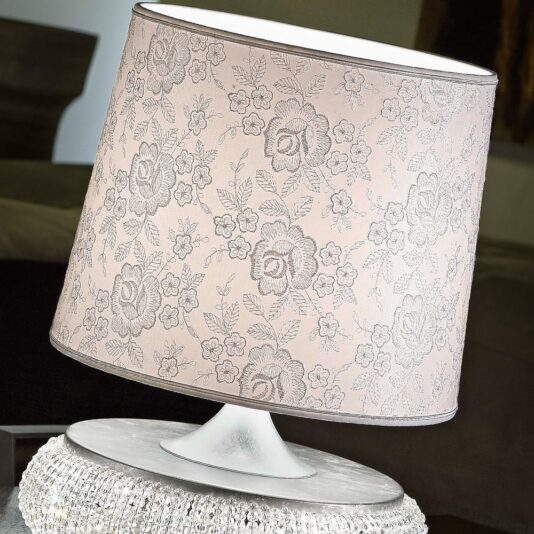A close-up of the Glamorous Chrome Crystal Table Lamp reveals a lampshade with an intricate floral pattern in shades of gray on a light pink background. The lampshade is perched on a decorative chrome base embellished with small crystals. The background is blurred, offering glimpses of a dark room.