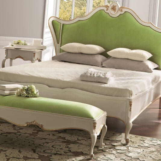 Elegant bedroom with a vintage white and green bed featuring a high, upholstered headboard. The room includes a matching White Lacquered Louis Reproduction Upholstered Bench at the foot of the bed, two stacks of folded towels, and small flower arrangements. Light streams in through large windows.