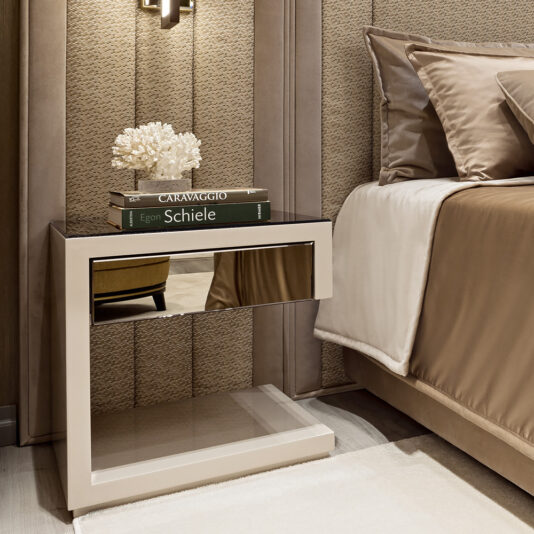 A High-End Contemporary Italian Designer Bedside Table with a mirrored drawer displays two art books and a white coral décor beside a neatly made bed with taupe and beige bedding in a stylish bedroom.