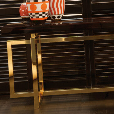 High End Contemporary Italian Rectangular Console A High-End Contemporary Italian Rectangular Console with dark wooden surfaces and golden metal legs elegantly showcases two tall, colorful vases adorned with geometric patterns in black, white, and orange alongside a small bowl. The backdrop features dark, partially open blinds that reveal a structured and modern room design.