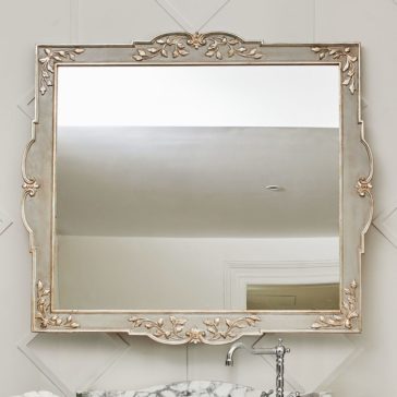 High End Louis XVI Inspired Marble Bathroom Vanity Unit A rectangular wall-mounted mirror with an ornate, vintage-style silver and gold frame featuring intricate floral and vine details reflects a portion of the white ceiling and part of a doorway. Below the mirror is a High-End Louis XVI Inspired Marble Bathroom Vanity Unit with silver faucet fixtures.