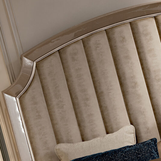 High End Italian Upholstered Storage Bed A close-up of the High End Italian Upholstered Storage Bed's elegant, high-end headboard showcases its glossy, curved edge. The headboard features vertical, plush panels in a beige color with light framing and a blue cushion partially visible in front of it—a true embodiment of refined luxury.