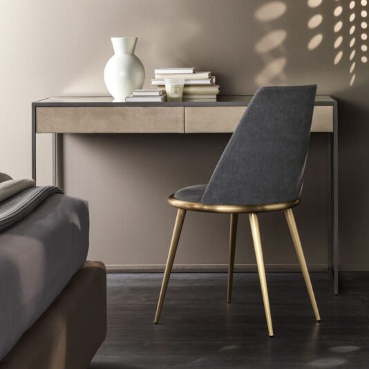 A modern bedroom corner features the High End Modern Italian Faux Nubuck Desk With Drawers, stacked books, a white vase, and a gray chair with gold legs. Sunlight projects leaf-shaped shadows above the desk, with part of a bed in the foreground.