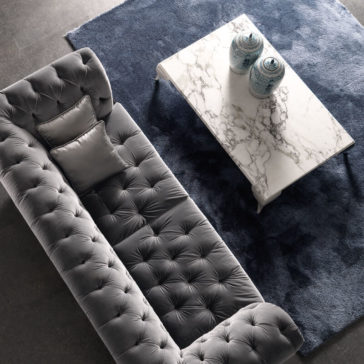 A top-down view of a modern living room showcases a long, tufted gray sofa paired with a dark blue rug. The High End Rectangular Marble Coffee Table rests elegantly on the rug, adorned with two blue and white porcelain vases. A silver satin pillow is stylishly placed on the sofa.